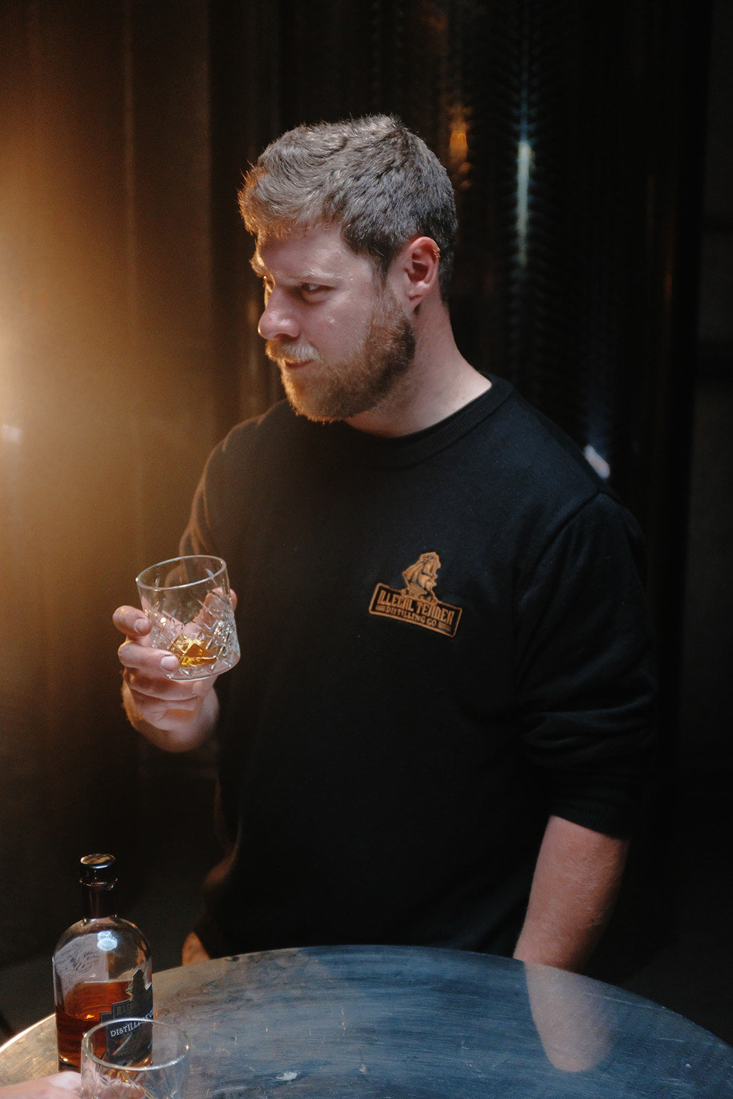 Left side view of a man holding a rum glass wearing a black Illegal Tender Rum Co sweatshirt.